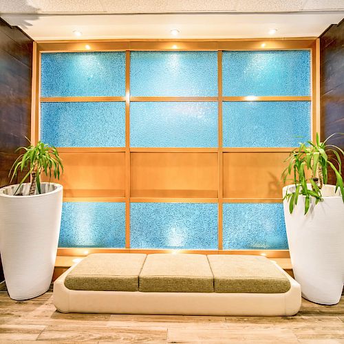 The image shows a modern indoor sitting area with a cushioned bench, two large potted plants, and a wall with blue glass panels framed in wood.