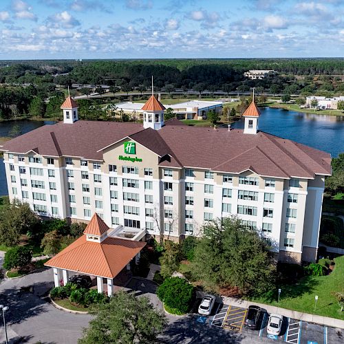 The image shows a multi-story hotel building with a red roof, located beside a body of water, surrounded by greenery, and parking lots.