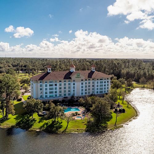 A large hotel with a red-roofed building is set by a scenic lake, surrounded by lush greenery and trees under a partly cloudy sky.