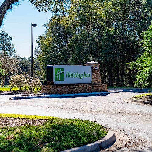 The image shows a Holiday Inn sign at an entrance, surrounded by trees and greenery, with clear skies in the background.