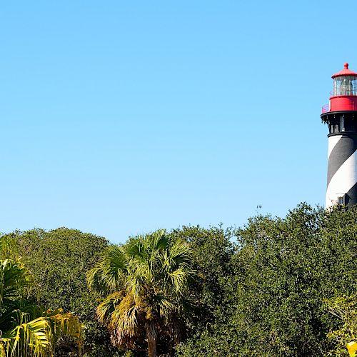 A lighthouse with black and white stripes and a red top is visible above green foliage under a clear blue sky.