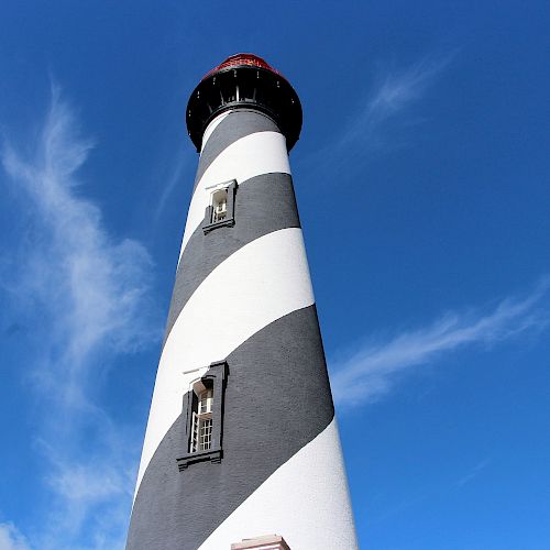 A tall lighthouse with a black and white spiral pattern against a blue sky with wispy clouds.