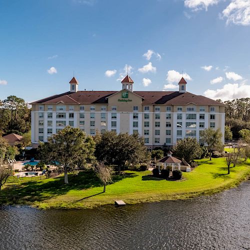 This image shows a large hotel building with several floors, adjacent to a body of water. The green lawn and trees surround the area, ending the sentence.