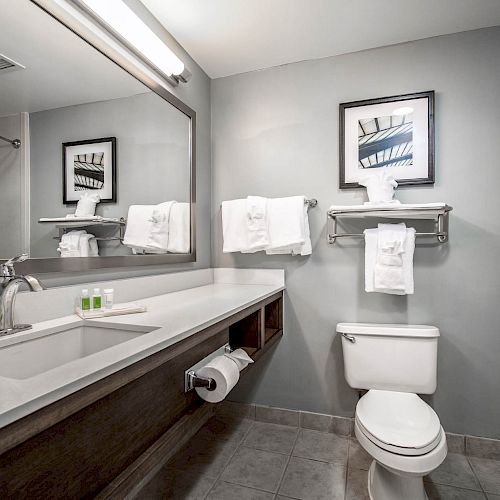 This image shows a modern bathroom with a sink, a large mirror, towels, a toilet, a bathtub, toiletries, and a wall art piece above the towel rack.