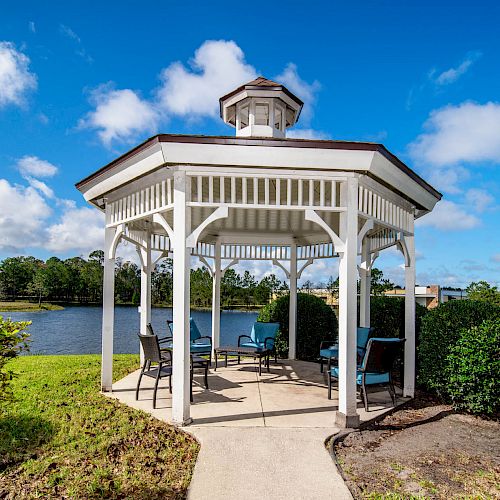 This image shows a white gazebo by a lake with outdoor seating, surrounded by greenery and set against a bright blue sky with scattered clouds.