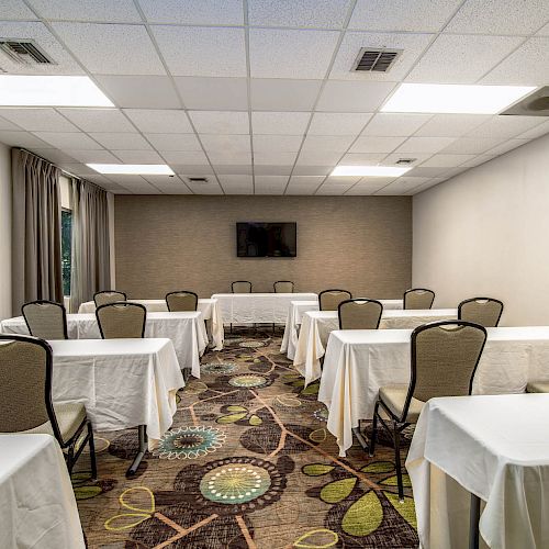 A small conference room with several tables and chairs, white tablecloths, and a TV mounted on the back wall.