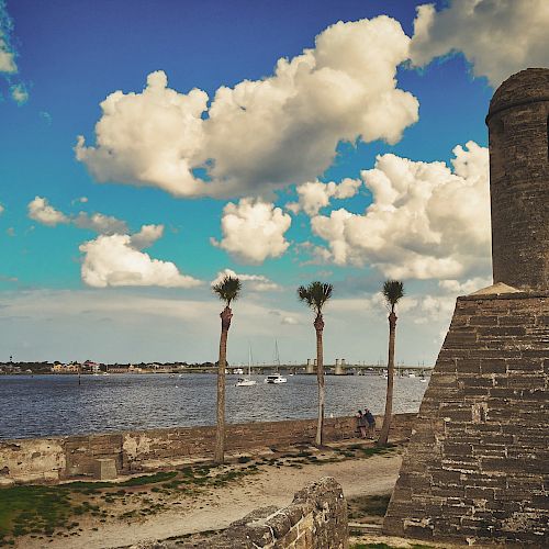 The image shows a historic stone fort by a body of water with palm trees and a partly cloudy sky in the background, creating a serene atmosphere.