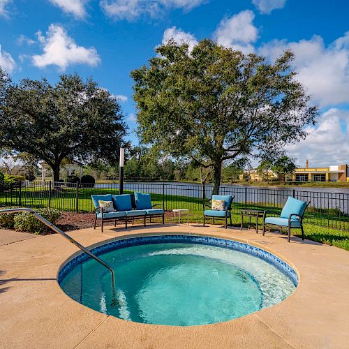 The image shows an inviting outdoor spa with a clear blue sky, surrounded by trees, lounge chairs, and a view of a lake in the background.