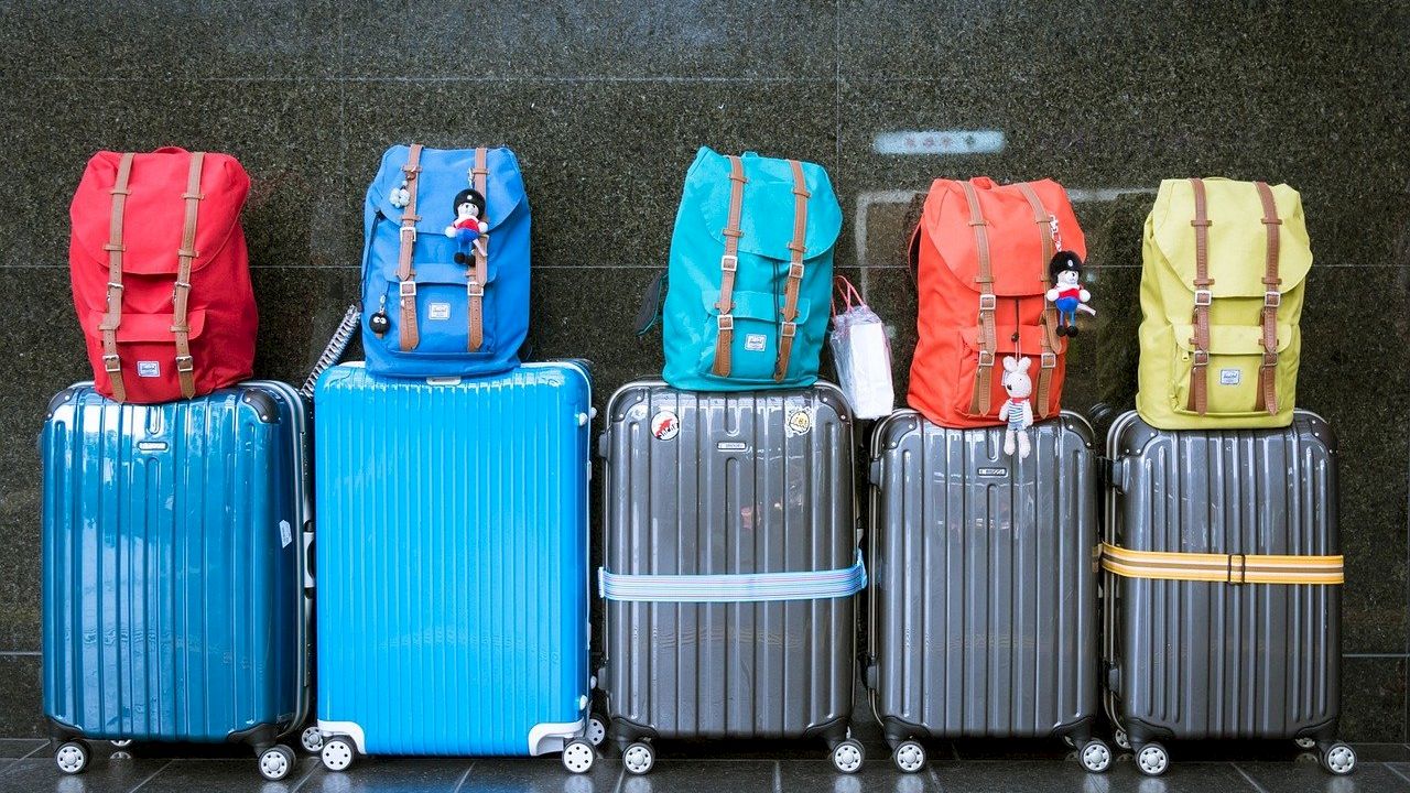 Several colorful suitcases and matching backpacks are lined up against a wall, each with small dolls attached to the front of the backpacks.
