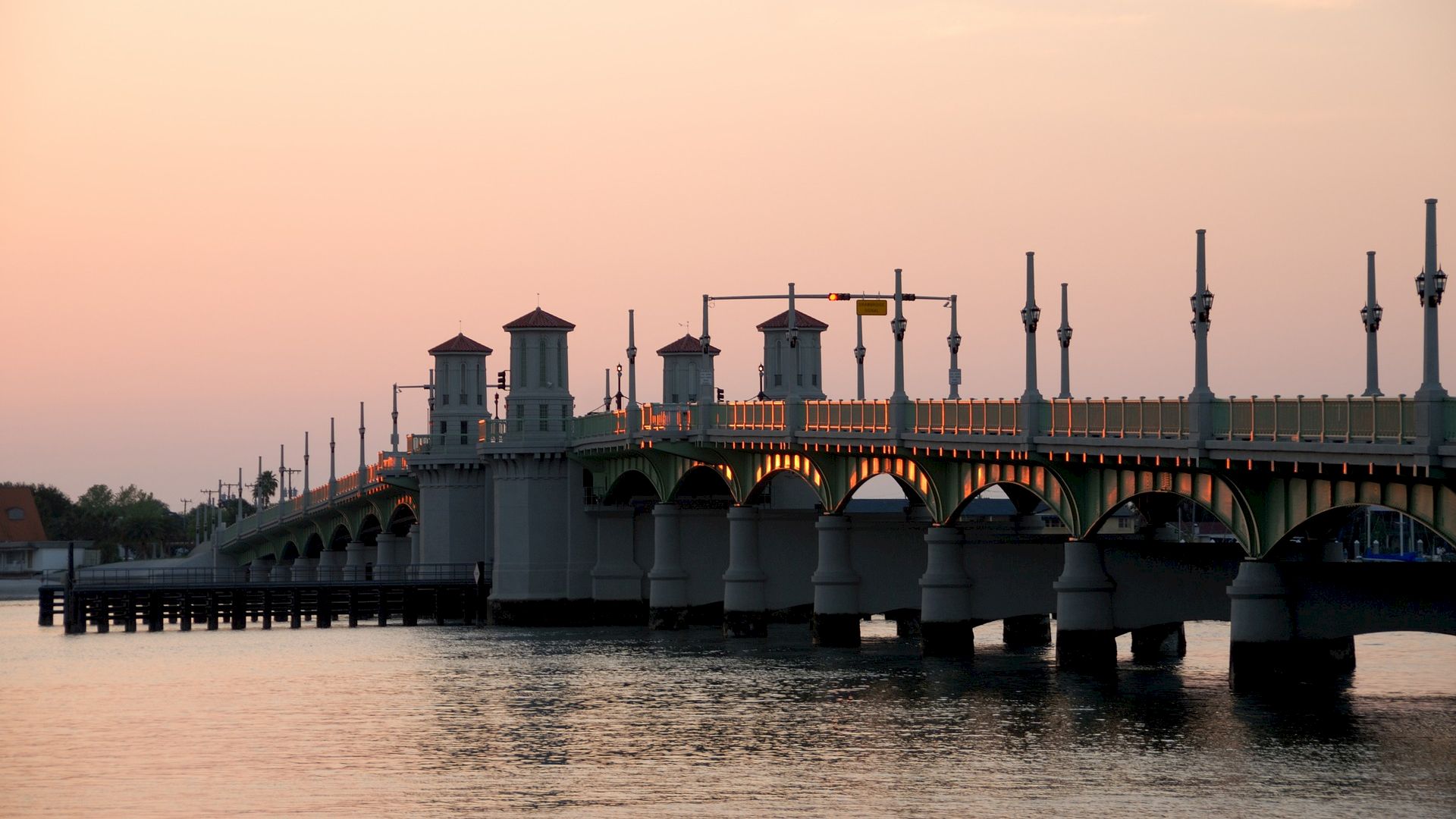 A bridge spans a body of water at sunset, with a warm sky and streetlights on the bridge silhouetted against the pinkish hue.