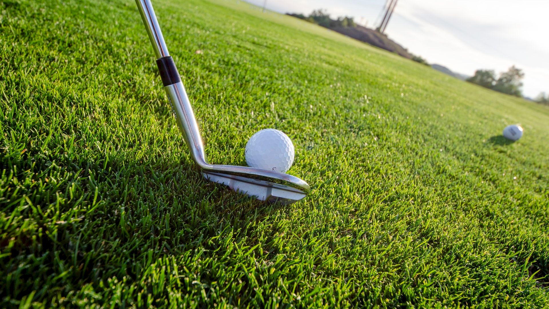 A golf club is positioned to hit a golf ball, with another golf ball visible in the background on a well-maintained green field under a clear sky.
