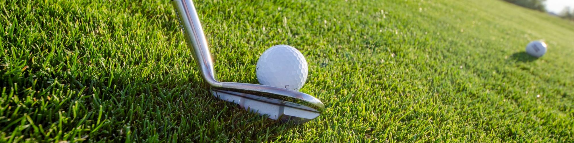 A golf club is positioned to hit a golf ball, with another golf ball visible in the background on a well-maintained green field under a clear sky.