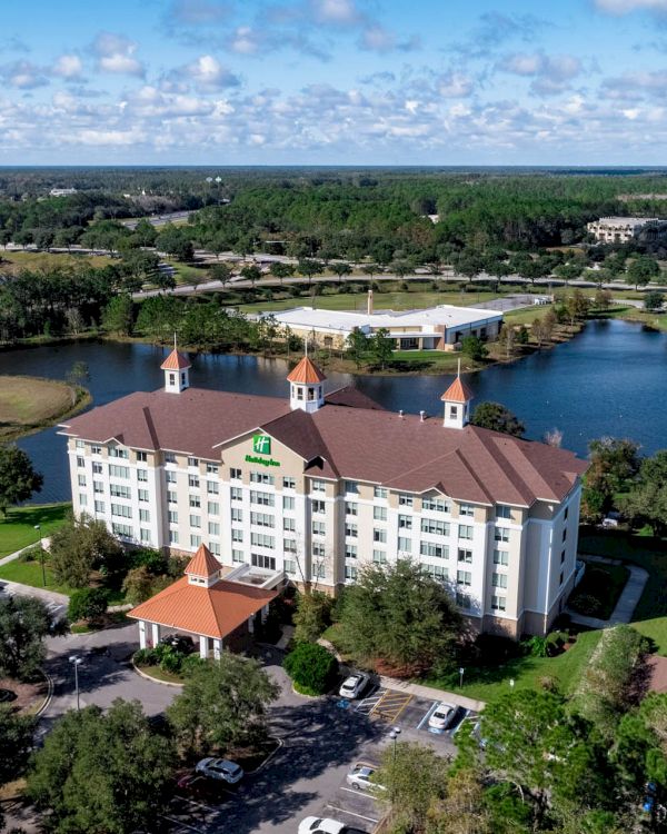 Aerial view of a large hotel building surrounded by trees, water, and parking areas, with a scenic landscape in the background.