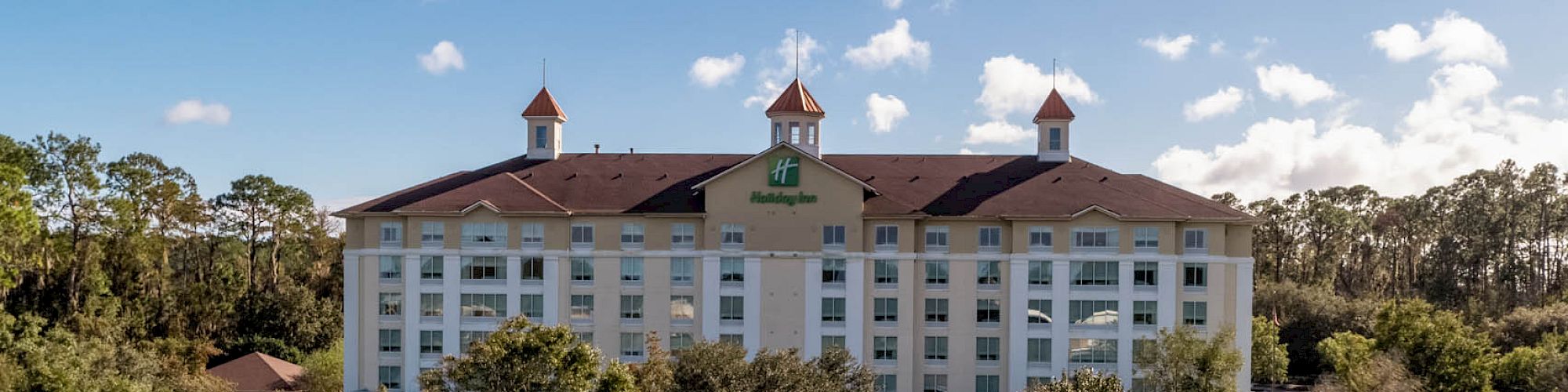 A large hotel near a body of water, surrounded by trees and greenery under a blue sky with clouds in the background.