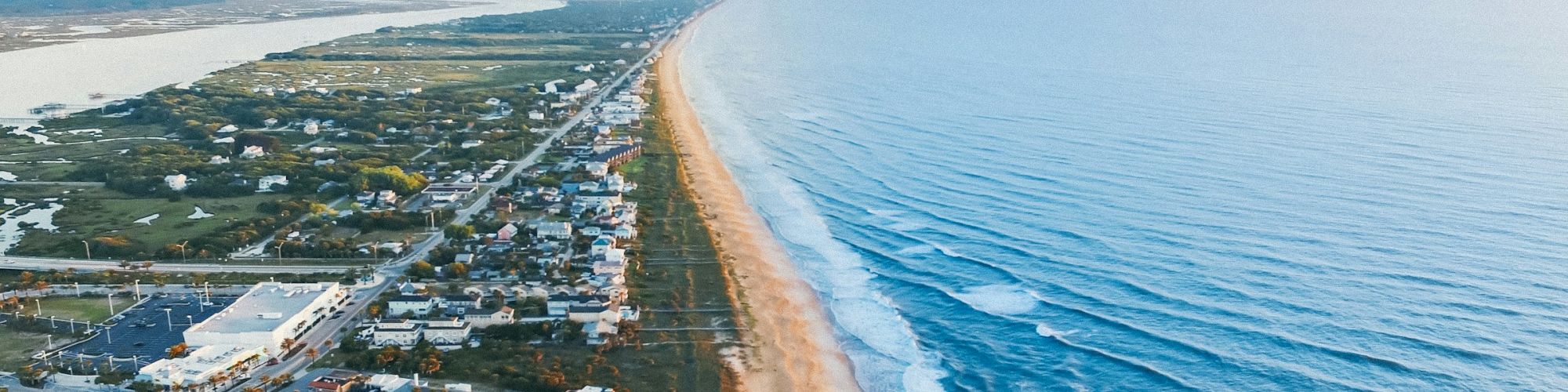 Aerial view of a coastal town with buildings, a sandy beach, and blue ocean waves extending into the horizon under a clear sky.