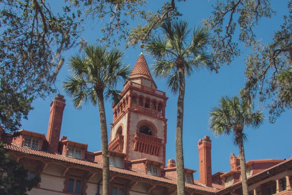 A historic building with a tall tower and red brick architecture, surrounded by palm trees and lush greenery under a clear blue sky.