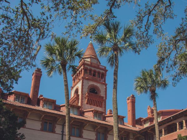 A historic building with a tall tower and red brick architecture, surrounded by palm trees and lush greenery under a clear blue sky.