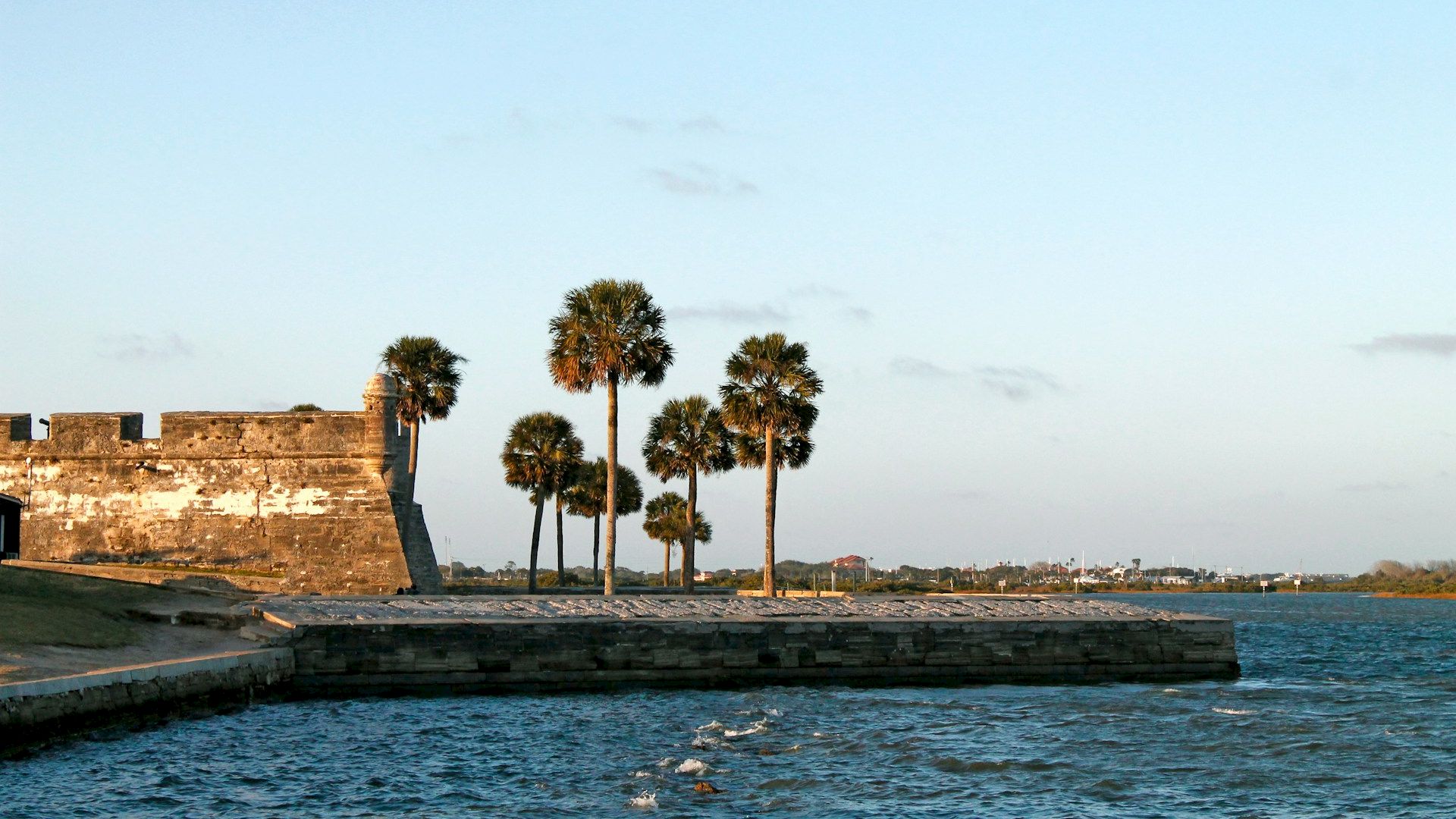 A waterfront scene with a stone fort, palm trees, and calm water under a clear sky.