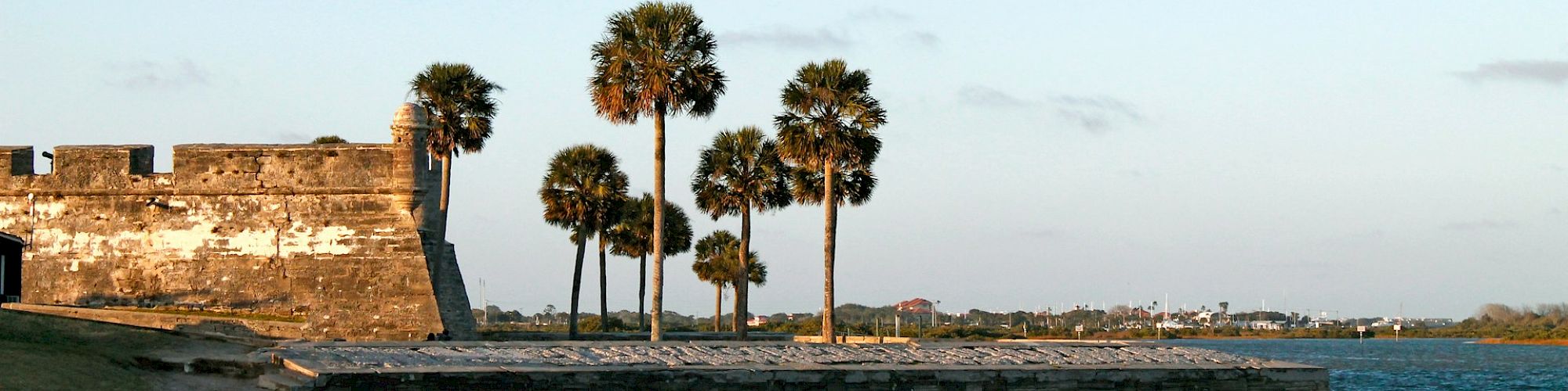 A waterfront scene with a stone fort, palm trees, and calm water under a clear sky.