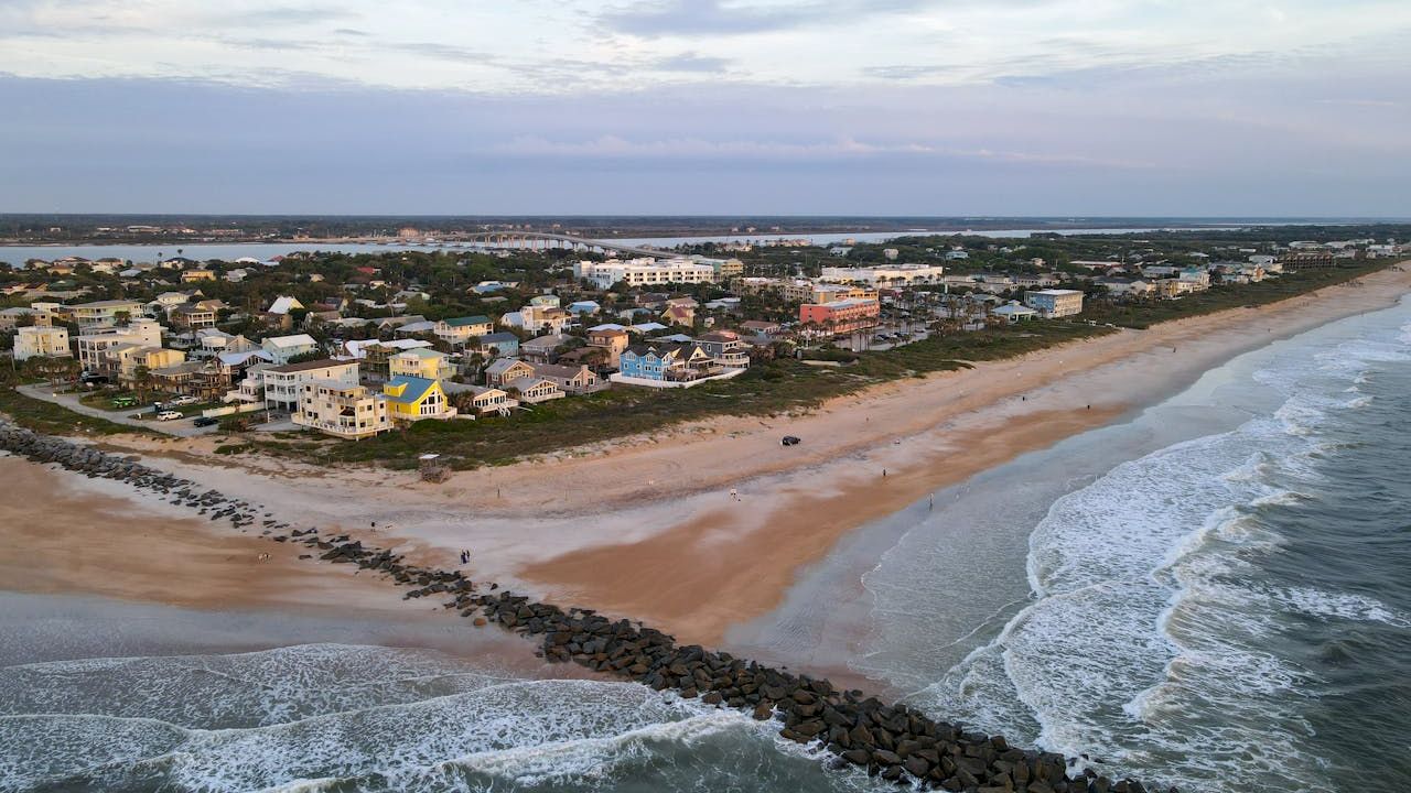A coastal town with a sandy beach, ocean waves, and a rocky jetty extending into the water, set under a partly cloudy sky.