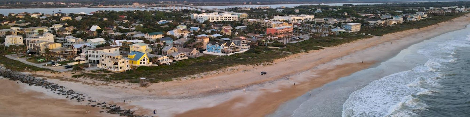 A coastal town with a sandy beach, ocean waves, and a rocky jetty extending into the water, set under a partly cloudy sky.
