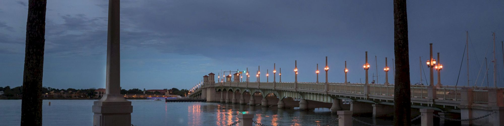 A lit bridge with ornate lamps spans a calm body of water under a cloudy evening sky, framed by a lamppost and palm tree.