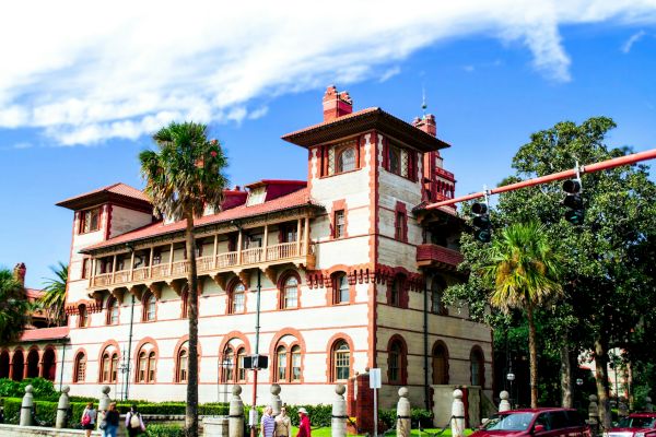 A historic building with red and white tones, featuring arches and palm trees in front, set against a blue sky with clouds.