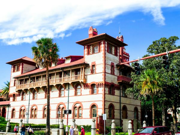 A historic building with red and white tones, featuring arches and palm trees in front, set against a blue sky with clouds.