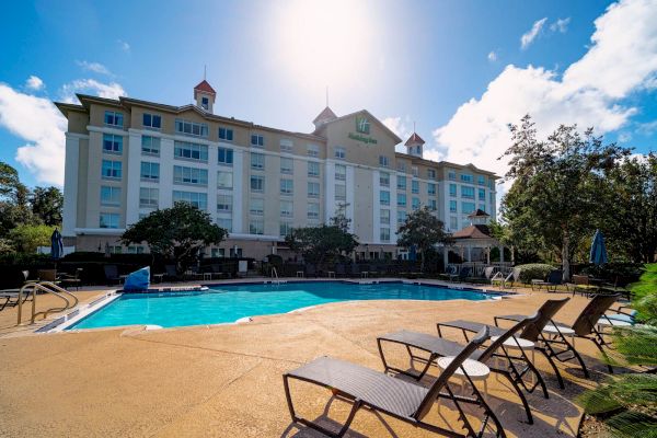 A hotel building with a swimming pool in front, surrounded by lounge chairs and trees under a bright sky.