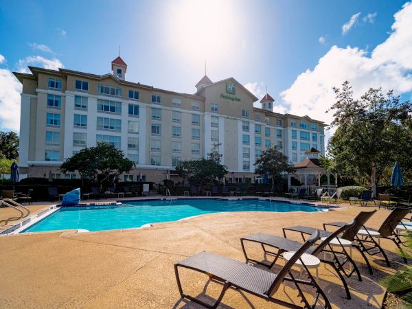 A hotel building with a swimming pool in front, surrounded by lounge chairs and trees under a bright sky.