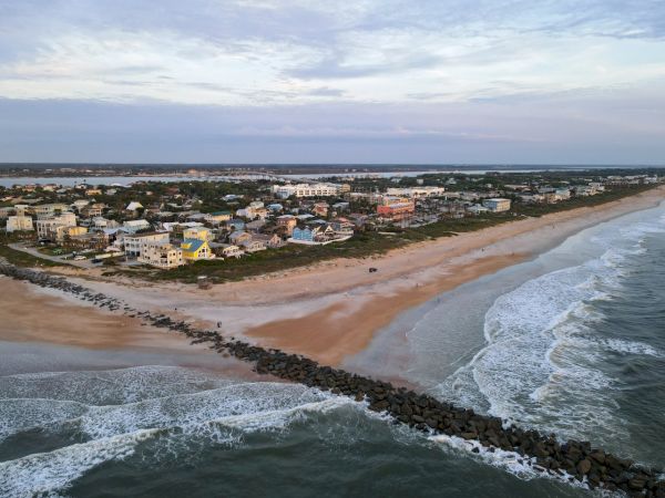 Aerial view of a coastal town with sandy beach, ocean waves, and a long stone jetty stretching into the water.