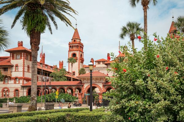 A historic building with red roofs and arched architecture, surrounded by lush greenery and palm trees, under a blue sky.