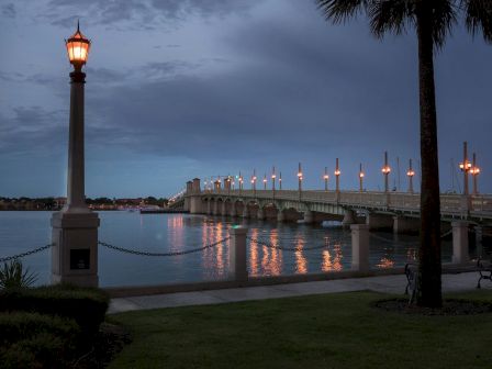 A scenic evening view of a lit-up bridge over calm water, with illuminated lampposts and reflections, framed by trees and a grassy area.