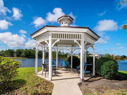 A white gazebo with chairs is by a lake under a blue sky with clouds, surrounded by greenery and trees, creating a peaceful setting.