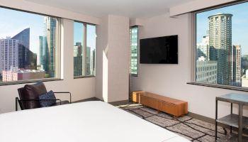 A modern bedroom with large windows offering a city view, featuring a bed, TV, armchair, and minimalistic decor.