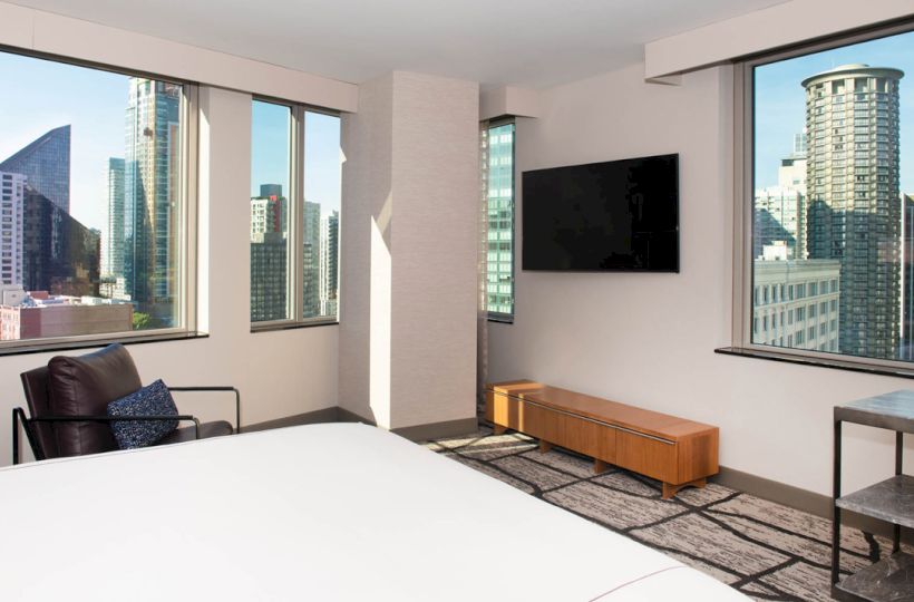 A modern bedroom with large windows offering a city view, featuring a bed, TV, armchair, and minimalistic decor.