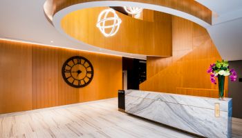 The image shows a modern lobby with a marble reception desk, a large wooden clock on the wall, and a spiral staircase.