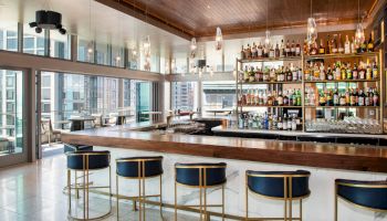 The image shows a modern bar with a wooden counter, blue and gold stools, and shelves stocked with various liquor bottles.