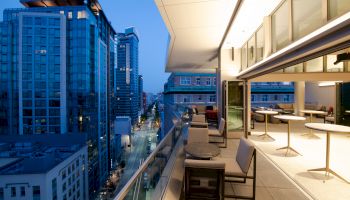 A modern high-rise balcony at dusk with outdoor seating overlooks a city street and skyline with lit-up buildings.