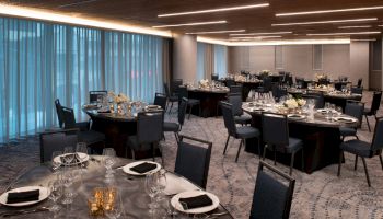 A dining room set up for an event, with round tables, black chairs, silverware, glasses, plates, and floral centerpieces.