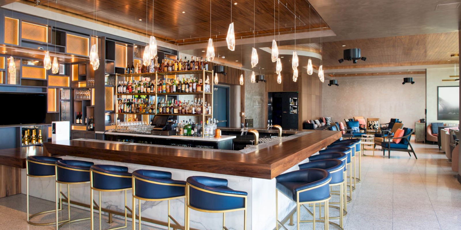 This image depicts a modern bar area with a central counter, blue bar stools, overhead pendant lights, and an extensive display of bottles.