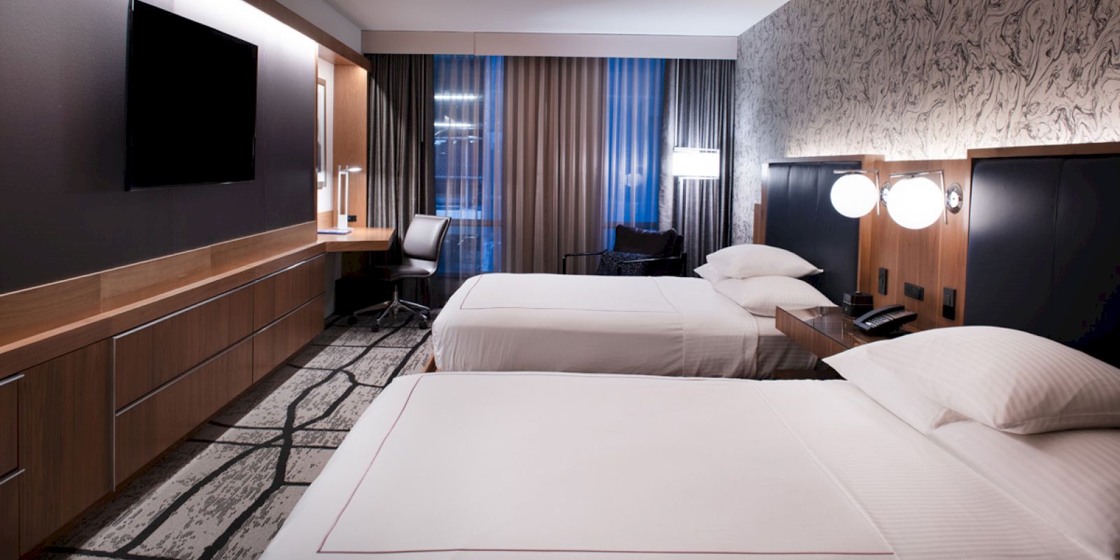 A modern hotel room with two beds, a flat-screen TV, a desk, and a chair; decorated with wood furniture and patterned surfaces ending the sentence.