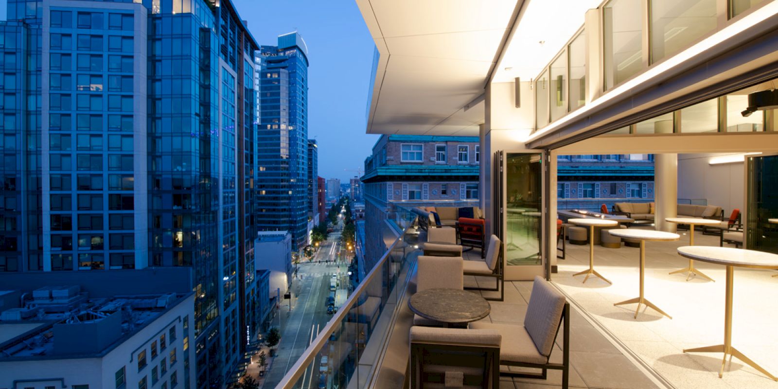 A balcony overlooking a cityscape at dusk, featuring outdoor seating and tables. The city has modern buildings and is illuminated by streetlights.