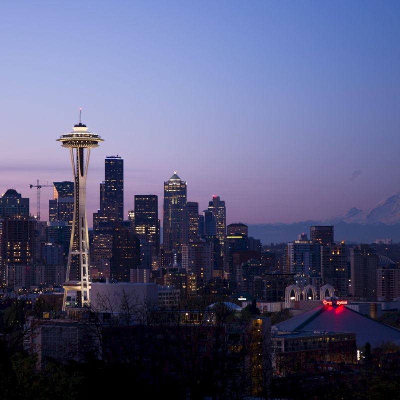 The image shows the Seattle skyline during twilight, featuring the Space Needle and Mount Rainier in the background, with the city's buildings illuminated.
