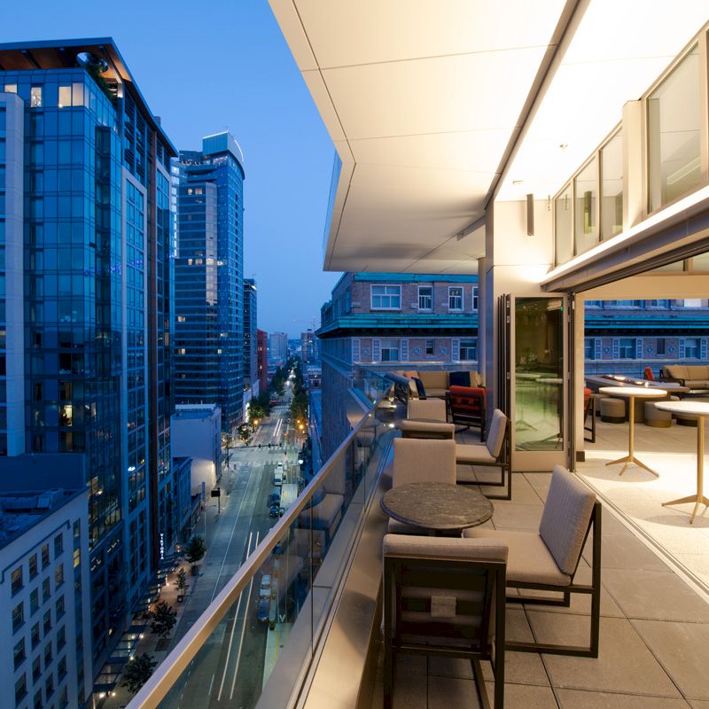 A modern urban balcony at twilight with seating areas overlooks a city street and high-rise buildings, reflecting a sleek and vibrant atmosphere.