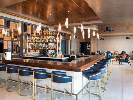 A modern bar with a wooden countertop surrounded by blue chairs, pendant lighting, a variety of bottles, and a seating area in the background.