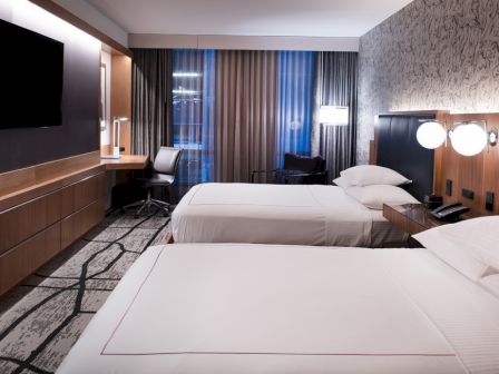 The image shows a modern hotel room with two neatly made beds, a flat-screen TV on the wall, a desk, lamps, and large windows with drapes.
