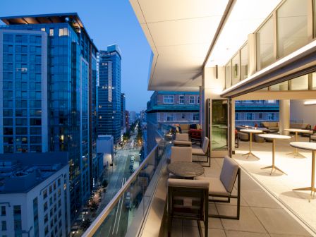 A modern cityscape at dusk, featuring high-rise buildings and a stylish balcony with seating and tables overlooking the street below.