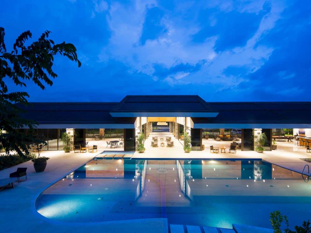 The image shows a luxurious, illuminated pool area at night with a modern building and open-air seating in the background.