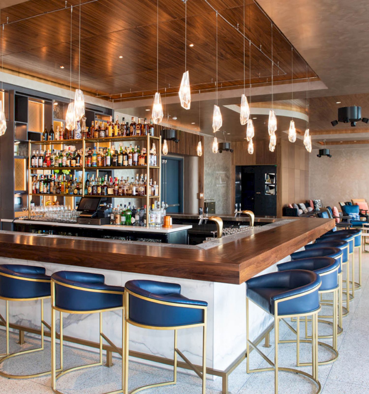 This image shows a modern bar with a wooden counter, blue stools, hanging lights, stocked shelves, and a cozy seating area in the background.